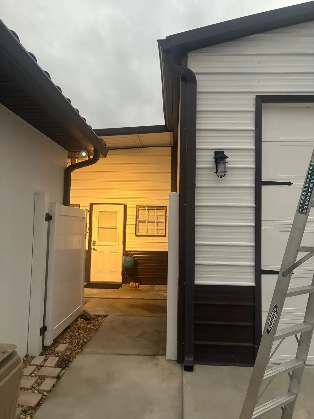 Brown gutter and downspout running down a metal building beside a white vinyl gate